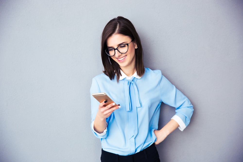 Smiling businesswoman using smartphone over gray background. Wearing in blue shirt and glasses.-1