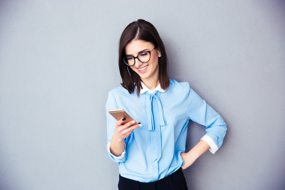 Smiling businesswoman using smartphone over gray background. Wearing in blue shirt and glasses.-1